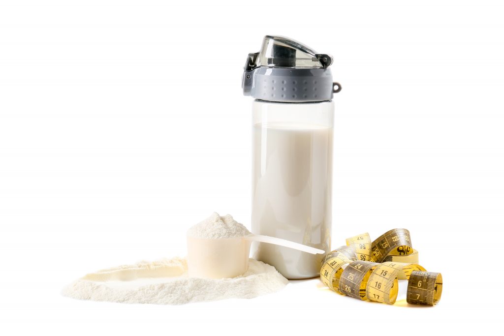 Bottle of protein shake with measuring tape on white background with a spoon full overflowing whey protein powder representing the popular appeal of Impact Whey.