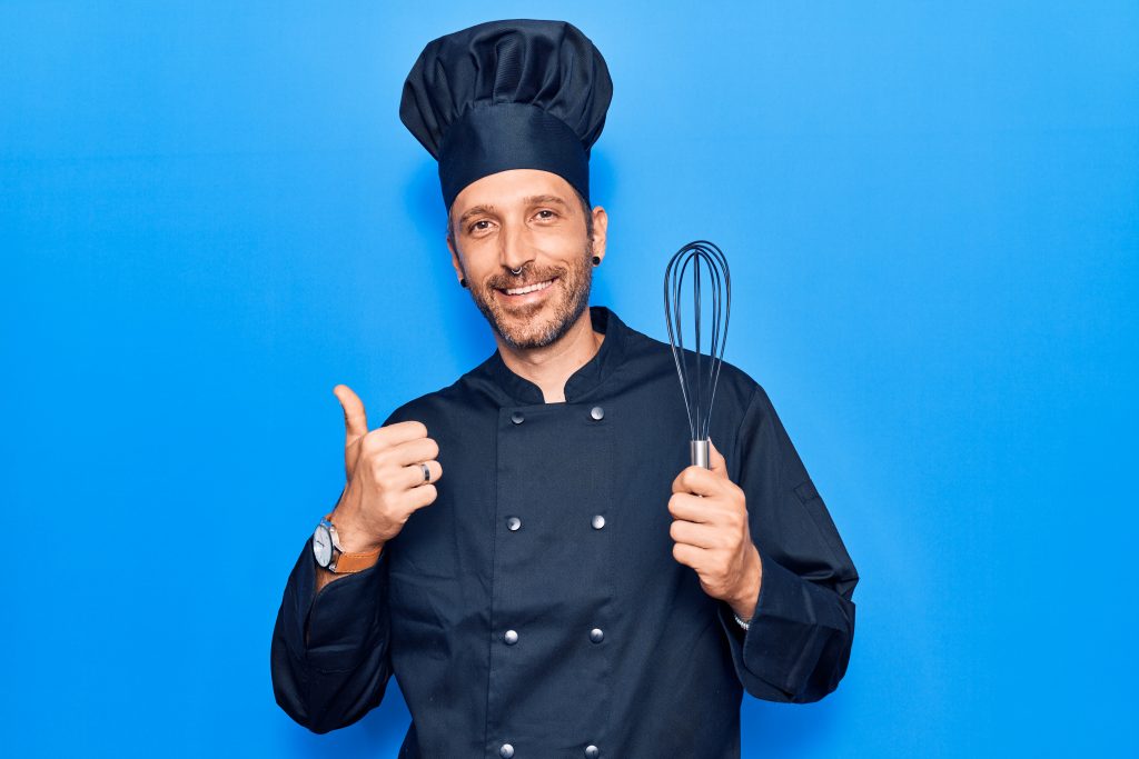 Man wearing blue chef clothes and a hat doing a thumbs up with a hand whisk