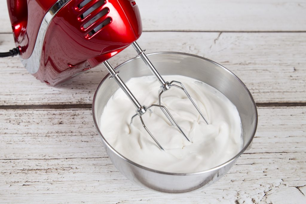 A red colored hand mixer with beater rods resting in a bowl of whipped cream.